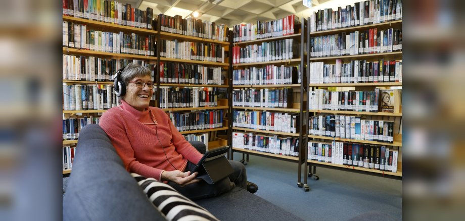 Frau mit Tablet und Kopfhörern sitzt in der Stadtbücherei vor Bücherregalen